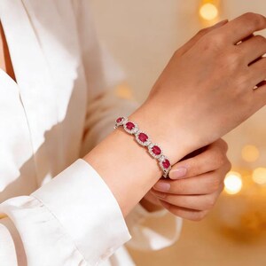 May include: A silver bracelet featuring oval-cut red gemstones, each surrounded by smaller, clear stones. The bracelet is worn on a wrist, showcasing its design and sparkle. The jewelry is set against a soft, neutral background.