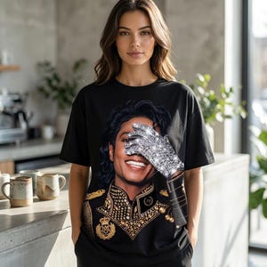 Könnte beinhalten: Schwarzes T-Shirt mit einem Porträt von Michael Jackson, dessen Gesicht teilweise von einem funkelnden Handschuh bedeckt ist. Das Shirt zeigt seine Jacke im Militärstil mit goldenen Details. Die Person steht in einer neutralen Umgebung mit einer hellen Arbeitsplatte und Hintergrund.