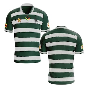 Puede incluir: Una camiseta de manga corta a rayas verdes y blancas con cuello en V. La camiseta presenta la bandera de Zimbabue en la manga y varios emblemas en el pecho, incluyendo una estrella y una cabra.