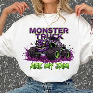 May include: White t-shirt featuring a graphic of a purple monster truck with green accents and large tires. The text "MONSTER TRUCK" is above the truck, and "ARE MY JAM" is below it, all in green and purple.