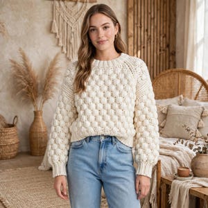 May include: A cream-colored long-sleeve sweater featuring a textured bobble knit pattern throughout the body and sleeves, paired with light blue denim jeans. The sweater has a round neckline and ribbed cuffs at the wrists and waistline.