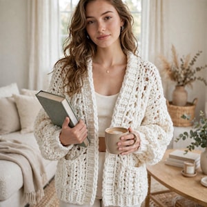 May include: An off-white chunky knit cardigan featuring an open front, long sleeves, and two front pockets. The garment is crafted with a thick, textured yarn in a loose, airy stitch pattern, providing a relaxed fit with a shawl collar neckline.