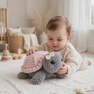 May include: A plush crochet turtle toy featuring a soft grey body and a textured light pink shell. The turtle has large black eyes, a small cream-colored bow on its head, and pink stitched cheeks. The yarn texture is thick and velvety in appearance.