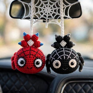 Puede incluir: Dos adornos de araña tejidos a ganchillo cuelgan de una telaraña blanca tejida. Una araña es roja con detalles de red negros y acentos azules, mientras que la otra es negra con detalles blancos. Ambas tienen ojos grandes y sonrisas.