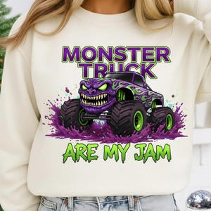 May include: Cream-colored sweatshirt featuring a purple monster truck graphic with green flames and accents. The truck has a menacing face and large tires, with the text "MONSTER TRUCK ARE MY JAM" in green and purple.