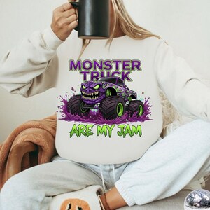 May include: White sweatshirt featuring a purple monster truck graphic with green accents and the text "MONSTER TRUCK ARE MY JAM". The design includes a monster truck with a menacing face and large tires.