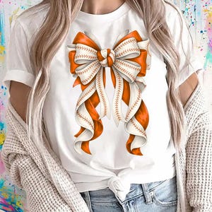 May include: White t-shirt with a large bow design. The bow features orange and white ribbons, with baseball-style stitching details. The design is centered on the shirt, creating a sporty and stylish look.
