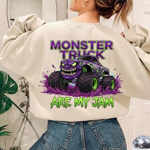 May include: Cream-colored sweatshirt featuring a purple and green monster truck graphic. The design includes the text "MONSTER TRUCK ARE MY JAM" in green and purple. The monster truck has a menacing face and large tires.