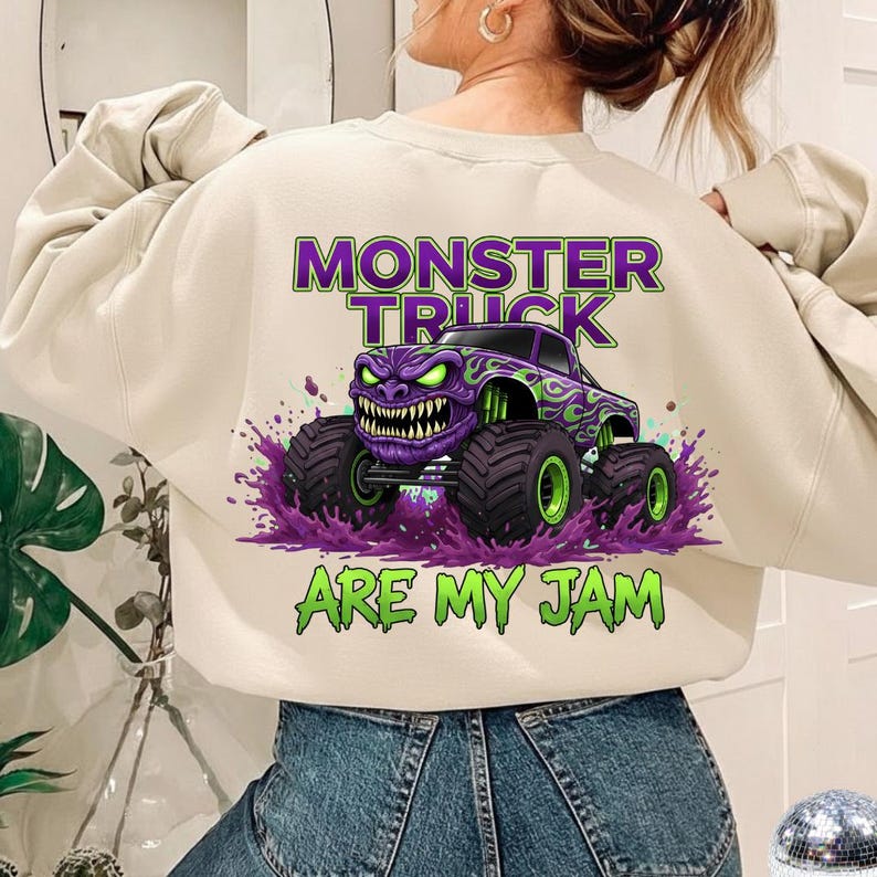 May include: Cream-colored sweatshirt featuring a purple and green monster truck graphic. The design includes the text "MONSTER TRUCK ARE MY JAM" in green and purple. The monster truck has a menacing face and large tires.