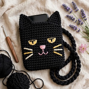 May include: A black crocheted crossbody bag shaped like a cat face with pointed ears, yellow embroidered eyes, a pink nose, and white whiskers. The bag features a thick braided black strap and contains a black smartphone inside the top opening.