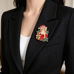 May include: A metal brooch featuring an orange tabby cat wearing a red Santa hat, peeking out from a red gift box with a gold ribbon. The design includes green holly leaves with white berries at the base, all finished with colourful enamel detailing.