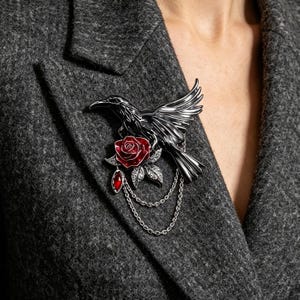 May include: A black metal raven brooch featuring a detailed bird in flight, a deep red enamel rose with silver-toned leaves, a dangling red teardrop-shaped gemstone, and two delicate silver-toned metal chains draped beneath the floral element.