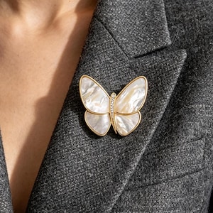 May include: A gold-toned butterfly brooch with iridescent white wings and a row of small clear stones on the body. The brooch is pinned to a gray blazer, showcasing the jewelry's design and color contrast.