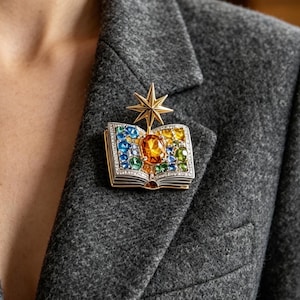 May include: A metallic brooch shaped like an open book with a gold star rising from the center. The pages are encrusted with small blue, green, and yellow gemstones, featuring a large oval orange stone in the middle of the open book design.