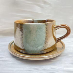 Può includere: Un set di tazza e piattino da caffè in ceramica. La tazza è cilindrica con un manico curvo e il piattino è rotondo. Il set presenta una miscela di colori, tra cui marrone, verde e beige, con una finitura craquelé.