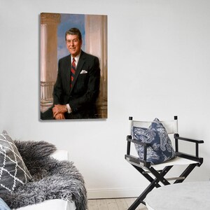 Puede incluir: Un retrato enmarcado de un hombre con un traje a rayas oscuras y una corbata a rayas rojas y azules. El retrato est&aacute; colgado en una pared blanca. Una silla de director con un coj&iacute;n azul estampado est&aacute; en primer plano.