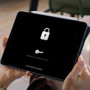 May include: A black tablet displaying a lock icon and a key icon, with the text "Drag the key to unlock our story" and "CLICK HERE TO OPEN." A white stylus rests on the tablet's edge.