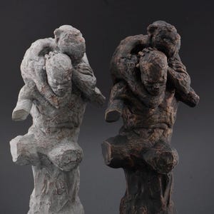 Puede incluir: Dos esculturas que representan a una figura cargando a otra sobre su espalda. Una escultura tiene un acabado gris claro con textura de piedra, mientras que la otra tiene un acabado oscuro, tono bronce y desgastado con detalles tallados visibles.