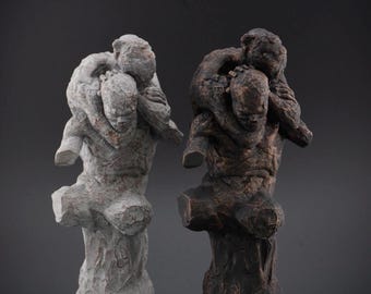 May include: Two sculptures depicting a figure carrying another figure on their back. One sculpture is finished in a light gray, stone-like texture, while the other is finished in a dark, bronze-toned, weathered texture with visible carved details.