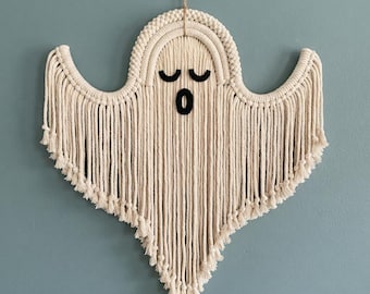May include: A macrame wall hanging shaped like a ghost, crafted from off-white cotton cord. The design features a rounded top, two outstretched arms, and a pointed bottom edge with fringe. Two black felt eyes and a black felt mouth are attached.