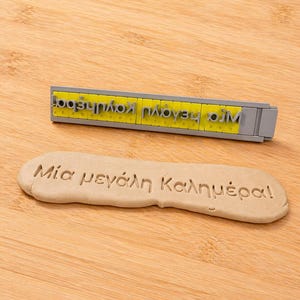 Puede incluir: Un juego de sellos gris y amarillo con texto griego, junto a un trozo de masa con la frase estampada "Mia megali Kalimera!" sobre una superficie de madera. El juego de sellos está diseñado para imprimir texto.
