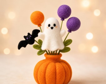 May include: A needle felted decoration featuring a white ghost, a black bat, and three round balloons in orange and purple, all emerging from a bright orange pumpkin-shaped base with green felt leaves at the base of the figures.
