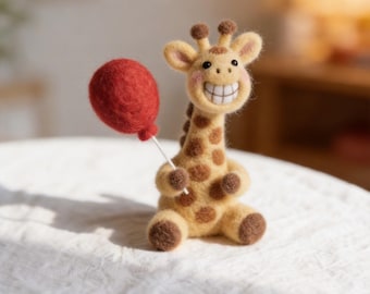 May include: A handmade felt giraffe with a big smile, holding a red balloon. The giraffe is tan with brown spots and a light brown mane. The balloon is round and attached to a white stick. The giraffe is sitting on a white surface.