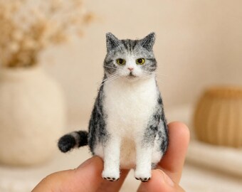 May include: A small needle-felted cat figurine featuring a grey and white tabby coat pattern. The cat has pointed ears, yellow eyes, long white whiskers, and a white chest and paws. The textured wool surface shows detailed grey markings.