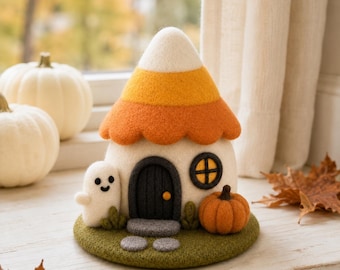 May include: A needle-felted candy corn house figurine featuring a white, orange, and yellow conical roof, a dark gray arched door, a small round window, a white ghost figure, and an orange pumpkin, all set on a circular green felt base.
