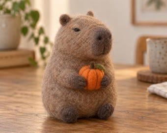 May include: A needle-felted capybara figurine made of light brown wool, featuring dark brown paws and facial details. The capybara is sitting upright and holding a small, bright orange pumpkin with a green stem between its front paws.