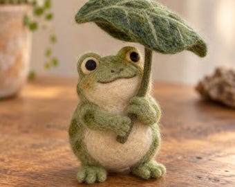 May include: A needle-felted frog figurine with a light green and cream-colored body, dark green spots, and large black eyes. The frog holds a large, textured green leaf over its head like an umbrella, with its front limbs wrapped around the stem.