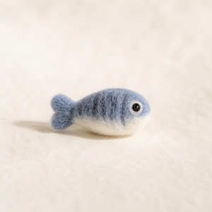 Pode incluir: Uma pequena figura de peixe feita com feltro de agulha e fibras de lã macia. O peixe apresenta um corpo arredondado com um padrão de cor degradê, que transita de um azul poeirento na parte superior para um branco limpo na parte inferior.