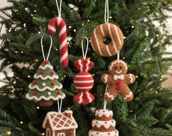 May include: A set of seven felt Christmas ornaments hanging on a tree, including a red and white candy cane, a brown donut, a green Christmas tree, a red and white wrapped candy, a gingerbread person, a gingerbread house, and a cake.