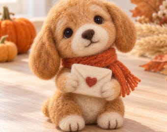 May include: A needle-felted tan puppy figurine with floppy ears, wearing a textured orange scarf and holding a small cream-colored envelope featuring a single red heart detail on the front, crafted with a soft, fuzzy wool texture.