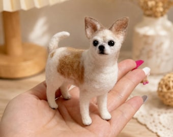 May include: A small needle-felted chihuahua figurine with white and tan fur, pointed ears, and black bead eyes. The dog is standing in a alert pose with its tail curled upward, showcasing a soft, textured wool finish and detailed facial features.