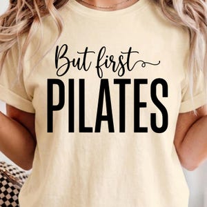 May include: A pale yellow short-sleeve t-shirt featuring black text printed on the front. The text reads But first in a cursive script font, followed by the word PILATES in a large, bold, sans-serif capital letter font.