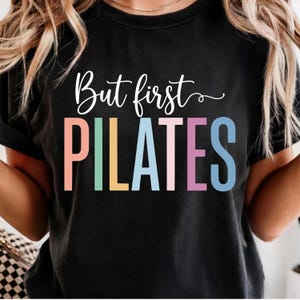 May include: A black short-sleeved t-shirt featuring the white cursive text But first above the word PILATES in large, block capital letters. Each letter of the word PILATES is coloured in a different pastel shade, including peach, mint, yellow, blue, pink, and light blue.
