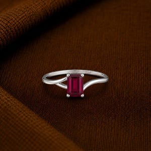 May include: A silver-toned ring featuring a single emerald-cut deep red gemstone set in a four-prong setting. The band has a bypass design where the two ends of the metal shank curve around the central stone, creating a sleek and polished appearance.