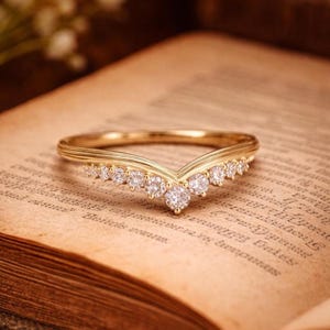 Könnte beinhalten: Ein goldener V-förmiger Ring mit einer Reihe kleiner, runder, klarer Steine, die entlang des chevronartigen Bandes gefasst sind. Das Metall hat eine polierte Oberfläche und die Steine sind in einem symmetrischen Muster angeordnet.