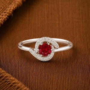 Puede incluir: Un anillo de plata con una piedra preciosa redonda de color rojo intenso en el centro, rodeada por un engaste en espiral incrustado con pequeñas piedras blancas transparentes que se extienden a lo largo del aro, con acabado brillante.
