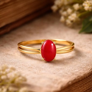 Può includere: Un anello d'oro con una vibrante pietra preziosa rossa ovale. L'anello presenta un design a fascia divisa ed è posto su uno sfondo neutro, evidenziando il colore e i dettagli del gioiello. L'anello è un gioiello.