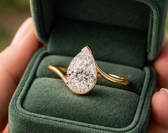 May include: A gold ring featuring a single pear-shaped faceted clear gemstone set in a smooth bezel setting. The band has a curved, asymmetrical design that tapers toward the stone. The ring is presented inside a dark green velvet box.