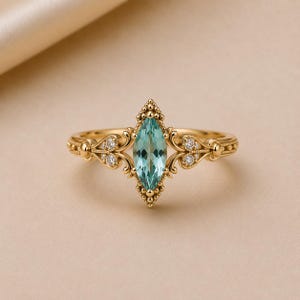 Op de afbeelding: Een gouden ring met een centrale lichtblauwe edelsteen in marquise-slijpvorm. De band is versierd met gedetailleerd metaalwerk en krulpatronen, geaccentueerd door vier kleine ronde witte steentjes in de symmetrische zijdetails.
