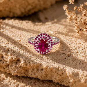 Peut inclure: Une bague en argent avec une pierre rouge centrale de taille ovale, entourée d'un double halo de petites pierres rouges rondes. L'anneau est également orné d'une rangée de petites pierres rouges sur chaque côté, créant un design rouge et argent.