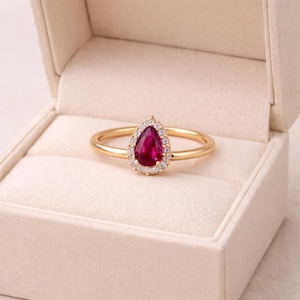 May include: A gold ring featuring a teardrop-shaped, deep red gemstone, encircled by small, clear stones. The ring is set in a gold band and displayed in a beige ring box. The ring's design is elegant and classic.