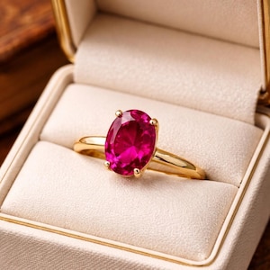 Peut inclure: Une bague en or avec une grosse pierre précieuse de couleur rubis, de forme ovale. La bague est sertie dans un sertissage classique à quatre griffes et est présentée dans un écrin blanc avec une bordure dorée. Le bracelet de la bague est en or poli simple.