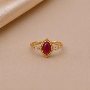 Könnte beinhalten: Ein goldener Ring mit einem zentralen, ovalen roten Edelstein, der von einem perlenartigen Metallrand umgeben ist. Das Band enthält zwei kleine, runde, klare Steine auf beiden Seiten der zentralen Fassung, was ein symmetrisches Design ergibt.