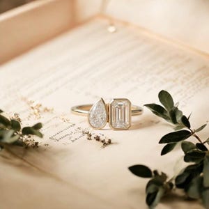 Peut inclure: Une bague en or ornée de deux pierres en diamant : une pierre en forme de poire et une pierre de taille émeraude. La bague est posée sur un papier vintage avec des accents floraux. C'est un bijou.