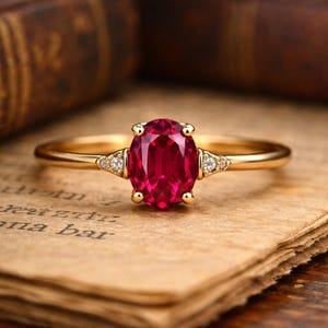 Pode incluir: Um anel de ouro com uma grande pedra de rubi oval como peça central. O anel tem pequenos diamantes em cada lado do rubi. O anel está sobre um fundo de papel vintage.