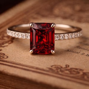 May include: A silver ring with a large, rectangular, deep red gemstone as the focal point. The band is adorned with small, clear gemstones. The ring is set against a vintage, brown background.